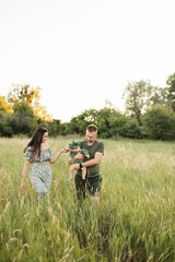 Wall Mural - Beautiful and happy caucasian parents playing with their six months old daughter walking on green field. Positive family of three enjoying leisure time on fresh air.