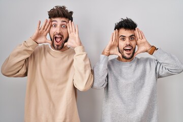 Wall Mural - Young homosexual couple standing over white background smiling cheerful playing peek a boo with hands showing face. surprised and exited