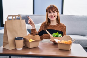 Wall Mural - Young beautiful woman eating delivery food at the living room smiling happy pointing with hand and finger to the side