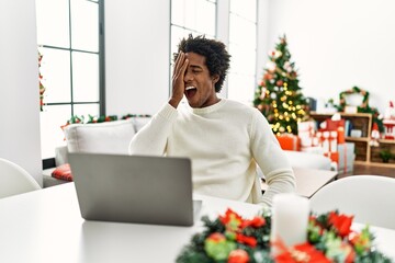 Poster - Young african american man using laptop sitting on the table by christmas tree covering one eye with hand, confident smile on face and surprise emotion.