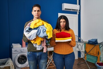 Sticker - Young couple holding laundry dirty and clean laundry skeptic and nervous, frowning upset because of problem. negative person.