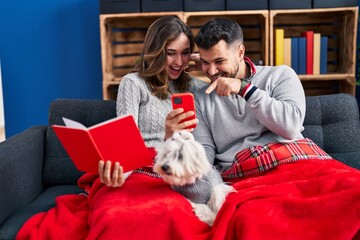  Man and woman reading book and using smartphone sitting on sofa with dog at home