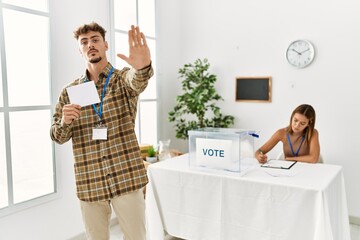 Poster - Young handsome man voting putting envelop in ballot box with open hand doing stop sign with serious and confident expression, defense gesture