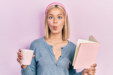 Poster - Beautiful blonde woman reading a book and drinking a cup of coffee making fish face with mouth and squinting eyes, crazy and comical.