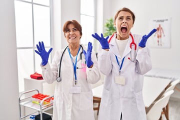 Two women wearing doctor uniform and stethoscope crazy and mad shouting and yelling with aggressive expression and arms raised. frustration concept.