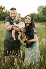 Wall Mural - Portrait of happy young parents embracing little cute daughter and their dog celebrate six months since the birth of a child while standing on the field on fresh air.