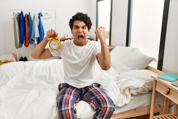 Wall Mural - Young hispanic man holding alarm clock sitting on the bedroom annoyed and frustrated shouting with anger, yelling crazy with anger and hand raised