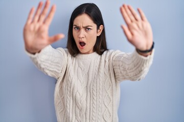 Poster - Young brunette woman standing over blue background doing stop gesture with hands palms, angry and frustration expression