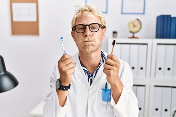 Poster - Young blond dentist man working at dentist clinic holding toothbrushes puffing cheeks with funny face. mouth inflated with air, catching air.