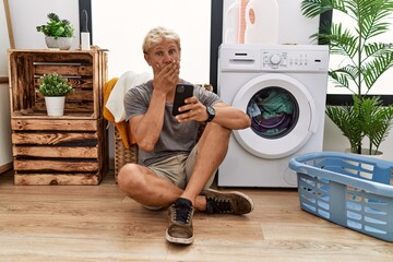 Poster - Young blond man doing laundry using smartphone shocked covering mouth with hands for mistake. secret concept.