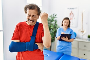 Wall Mural - Southeast asian man wearing wearing arm on sling at rehabilitation clinic annoyed and frustrated shouting with anger, yelling crazy with anger and hand raised