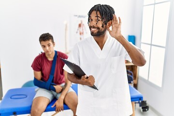 Poster - Young hispanic man working at pain recovery clinic with a man with broken arm smiling with hand over ear listening an hearing to rumor or gossip. deafness concept.