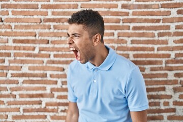 Poster - Brazilian young man standing over brick wall angry and mad screaming frustrated and furious, shouting with anger. rage and aggressive concept.