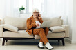 © Tatiana - a joyful elderly businesswoman is sitting on a cozy sofa in a bright apartment holding a laptop on her lap and gesturing with her hand while solving work issues on the phone
