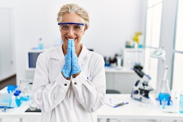 Wall Mural - Middle age blonde woman working at scientist laboratory praying with hands together asking for forgiveness smiling confident.