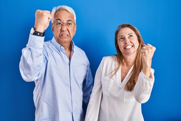 Sticker - Middle age hispanic couple standing over blue background angry and mad raising fist frustrated and furious while shouting with anger. rage and aggressive concept.