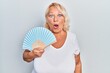 Middle age blonde woman waving hand fan cooling air in summer scared and amazed with open mouth for surprise, disbelief face