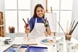 © Krakenimages.com - Young brunette woman at art studio with painted hands smiling looking to the camera showing fingers doing victory sign. number two.