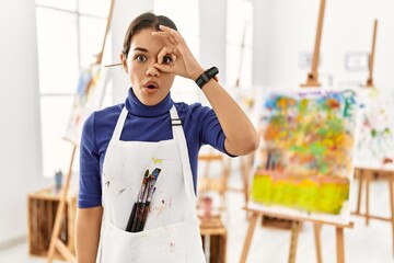 Canvas Print - Young brunette woman at art studio doing ok gesture shocked with surprised face, eye looking through fingers. unbelieving expression.