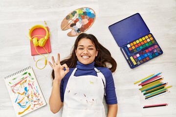 Sticker - Young brunette woman lying on the floor at art studio around artist things doing ok sign with fingers, smiling friendly gesturing excellent symbol