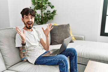 Poster - Hispanic man with beard sitting on the sofa celebrating crazy and amazed for success with arms raised and open eyes screaming excited. winner concept