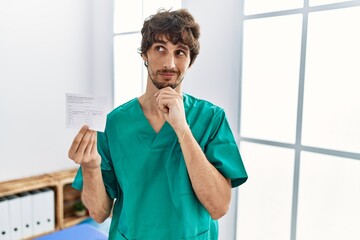 Sticker - Young hispanic doctor man holding covid record card with hand on chin thinking about question, pensive expression. smiling with thoughtful face. doubt concept.