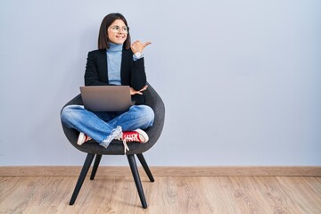 Young hispanic woman sitting on chair using computer laptop smiling with happy face looking and pointing to the side with thumb up.