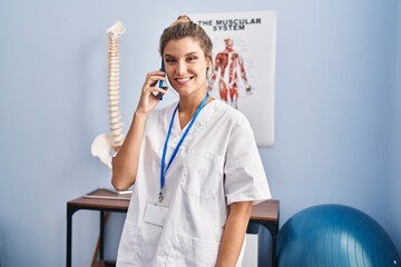 Poster - Young blonde woman wearing physiotherapist uniform talking on the smartphone at clinic