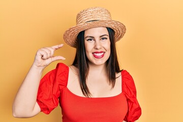 Poster - Young hispanic woman wearing summer hat smiling and confident gesturing with hand doing small size sign with fingers looking and the camera. measure concept.