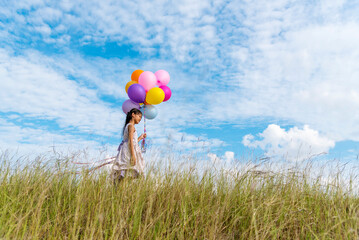 Wall Mural - Cheerful cute girl holding balloons running on green meadow white cloud and blue sky with happiness. Hands holding vibrant air balloons play on birthday party happy times summer on sunlight outdoor