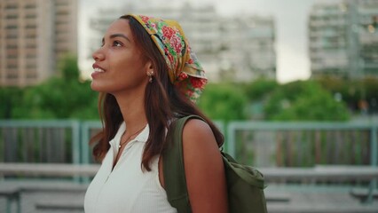 Wall Mural - Clouse-up, cute tanned woman with long brown hair in white top and yellow bandana with backpack on her shoulder and looks around the modern city. Camera moving around