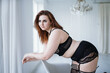 A plus size model in lacy black lingerie poses in a bright room with a trendy interior. Seductive chubby model in lingerie and harness