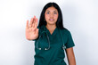 © Roquillo - Doctor hispanic woman wearing surgeon uniform over white wall doing stop sing with palm of the hand. Warning expression with negative and serious gesture on the face.