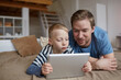© Mediaphotos - Serious blond boy lying on bed and using digital tablet while watching online movie with father