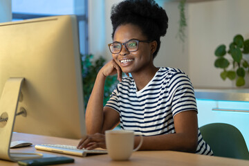 Joyful cheerful African American millennial woman looking at computer monitor, happy young black female sitting at table watching movie and drinking tea, enjoying leisure time in evening at home