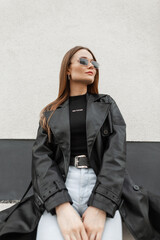 Beautiful street fashion girl in fashionable clothes with vintage gold sunglasses in a black leather coat with a black T-shirt and jeans sits in the city near the building