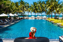 Luxury Beach Vacation In Tropical Beach Hotel. Tourist Woman In Red Dress Relax Near Blue Swimming Pool In Modern Resort. Female Traveler On Sea Vacation.