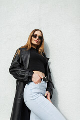 Fashionable beautiful young woman model with gold vintage sunglasses in a fashion long black leather coat with T-shirt and high-waisted jeans standing near the white wall on the street