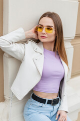 Wall Mural - Trendy hipster beautiful young woman model in fashionable bright casual street clothes with a leather jacket, purple T-shirt and jeans sits near a vintage building on the street