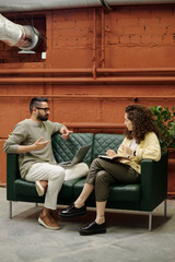 Wall Mural - Young confident businesswoman explaining something to male colleague while both sitting on green leather sofa and having meeting