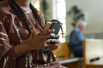 Hands of young black woman with Holy Bible and rosary beads with small wooden cross standing in church and praying after sermon