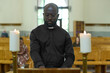 © pressmaster - African American priest in black shirt with clerical collar standing by wooden pulpit between two burning candles during church service