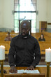 © pressmaster - Young African American man in black shirt with clerical collar reading one of four Gospels or some other book from Holy Bible during liturgy