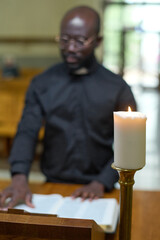 Wall Mural - White candle burning against young African American pastor reading Holy Bible while standing by pulpit in front of parishioners