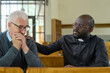 Young black man in shirt with clerical collar keeping hand on shoulder of senior male parishioner while comforting him and praying