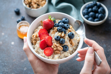 Wall Mural - Oatmeal porridge bowl with berry fruits in female hands, closeup view. Healthy vegetarian breakfast food