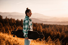 Woman Rides At Straight Road On Longboard At Sunset Time. Skater In Casual Wear Training On Board During Evening Sunset With Orange Light. Girl Hold Longboard In Hands