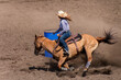 A Rodeo barrel racer is rounding a blue barrel. The cowgirl has a white hat, blue shirt and blue pants. The horse has a blond coat. The area is dirt.