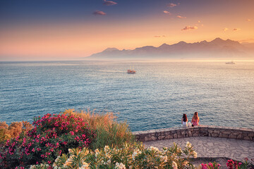 Wall Mural - Two girls friends are sitting resting and watching a beautiful bright sunset from an observation deck in the park. Wonderful mountains in the sunlight and ripples on the Mediterranean sea 