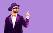 Smiling disco man in velvet jacket isolated on purple studio background point at blank empty copy space. Happy male entertainer in hat, glasses and suit recommend good sale deal or promotion.